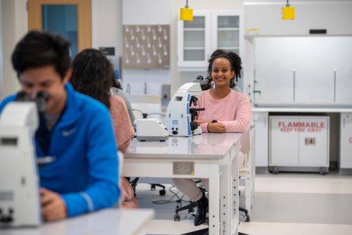 Student smiling at the camera while working in a lab, sitting in front of a miscroscope.